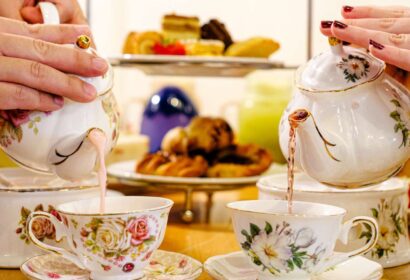 Two people pouring tea from floral teapots into matching cups, with a tiered tray of pastries in the background.