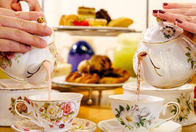 Two people pouring tea from floral teapots into matching cups, with a tiered tray of pastries in the background.