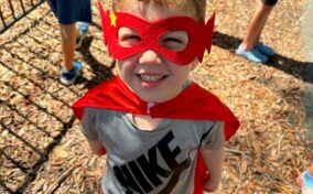 A child wearing a red superhero mask and cape smiles at the camera, standing on wood chips in a playground with other kids in the background.