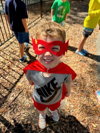 A child wearing a red superhero mask and cape smiles at the camera, standing on wood chips in a playground with other kids in the background.