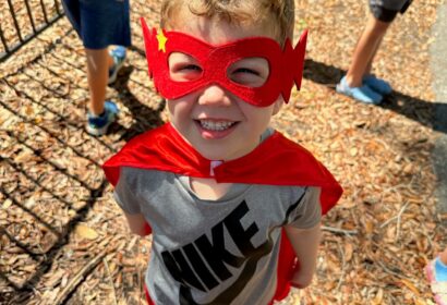 A child wearing a red superhero mask and cape smiles at the camera, standing on wood chips in a playground with other kids in the background.