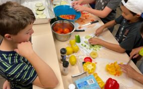 Children preparing vegetables on a counter, surrounded by sliced tomatoes, bell peppers, lemons, and spices. Various tools and cutting boards are visible.