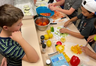 Children preparing vegetables on a counter, surrounded by sliced tomatoes, bell peppers, lemons, and spices. Various tools and cutting boards are visible.