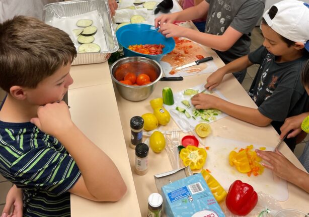 Children preparing vegetables on a counter, surrounded by sliced tomatoes, bell peppers, lemons, and spices. Various tools and cutting boards are visible.