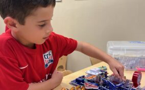 A young boy in a red shirt is assembling an electronic circuit on a table. Educational posters are visible on the wall behind him.