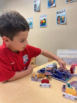 A young boy in a red shirt is assembling an electronic circuit on a table. Educational posters are visible on the wall behind him.