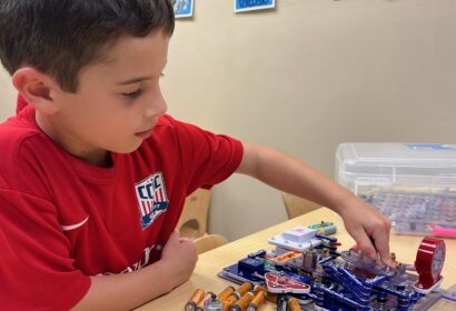 A young boy in a red shirt is assembling an electronic circuit on a table. Educational posters are visible on the wall behind him.