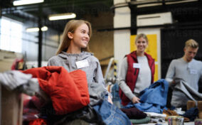 People sorting clothes at a charity event, with a woman in the foreground smiling beside a pile of clothing.