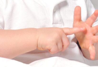 A baby in a white shirt is using their fingers to count, pointing with the right hand and holding up fingers on the left hand.