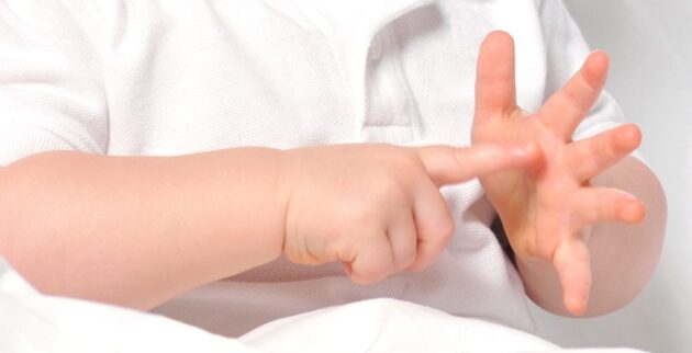 A baby in a white shirt is using their fingers to count, pointing with the right hand and holding up fingers on the left hand.