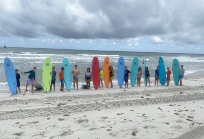 A group of people standing on a sandy beach, each holding a brightly colored surfboard, with the ocean and cloudy sky in the background.