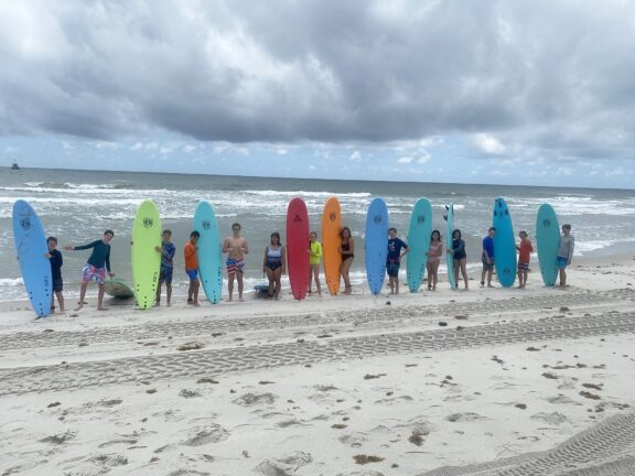 A group of people standing on a sandy beach, each holding a brightly colored surfboard, with the ocean and cloudy sky in the background.