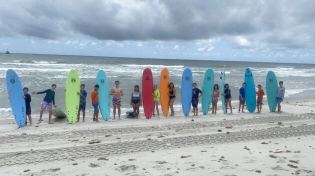 A group of people standing on a sandy beach, each holding a brightly colored surfboard, with the ocean and cloudy sky in the background.