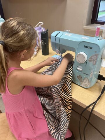 A young girl in a pink dress uses a sewing machine to sew a zebra-patterned fabric.