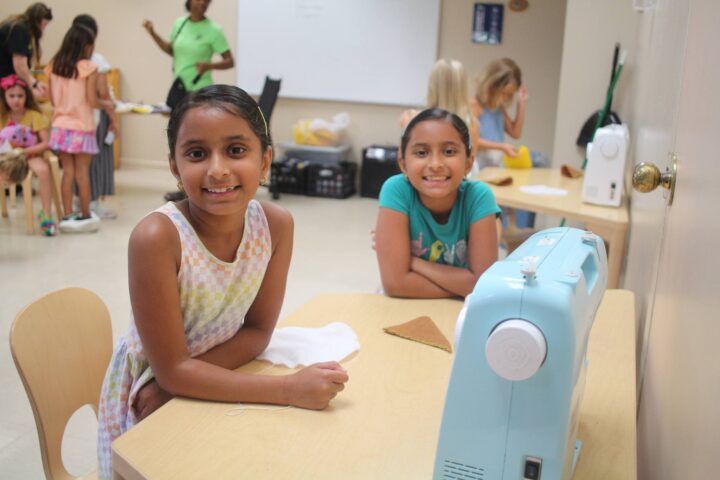 Two young girls smile at the camera while sitting at a table with a sewing machine. Other children and an adult are in the background.