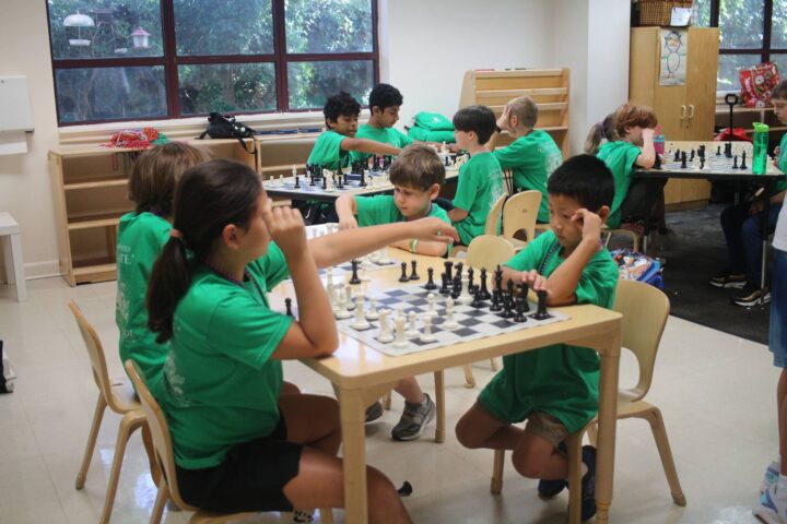 Children wearing green shirts are playing chess in a classroom setting, seated at tables with chess boards.