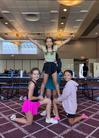 Three girls pose indoors with one lifted in the center, arms raised. They are in a room with tables and a patterned carpet.