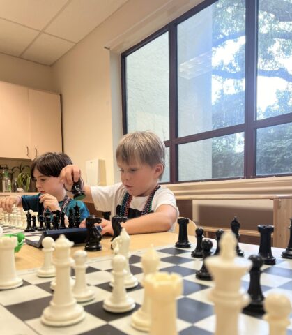 Two young boys focus on playing chess in a bright room with a large window. Chess pieces are visible on the board in the foreground.