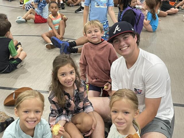 Group of children and a young adult sitting on a gym floor, smiling at the camera. Some children are eating snacks.