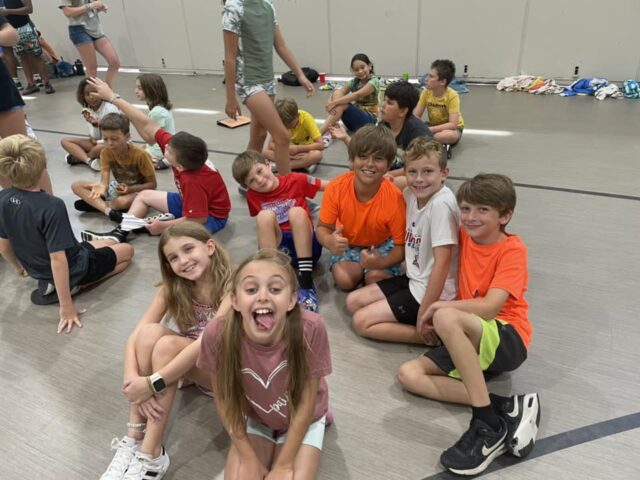 Children sitting on a gym floor, smiling and posing for the camera, with others interacting in the background.