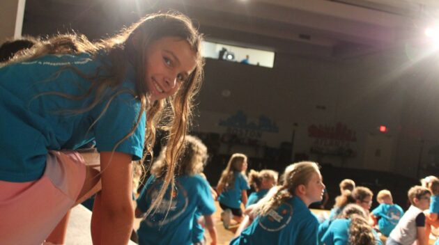 A group of children in blue shirts participate in an indoor activity, with one girl looking back at the camera. Stage lighting illuminates the scene.