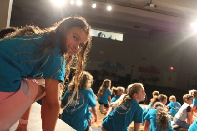 A group of children in blue shirts participate in an indoor activity, with one girl looking back at the camera. Stage lighting illuminates the scene.