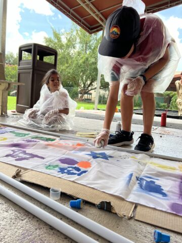 Two children in rain ponchos paint a long banner on the ground under a covered area, surrounded by art supplies.