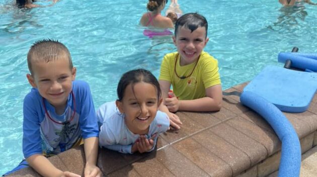 Three children smile at the edge of a pool, with others swimming and lounging in the background.