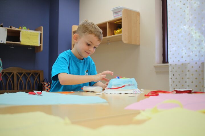 A young boy in a blue shirt sits at a table, focused on a crafting project. Surrounding him are colorful papers and tools in a room with shelves.