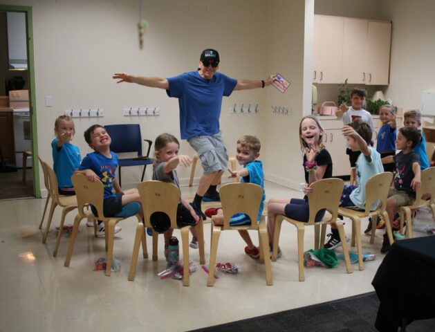 Man in a blue shirt and cap, mid-jump, surrounded by smiling children seated on chairs in a classroom setting.