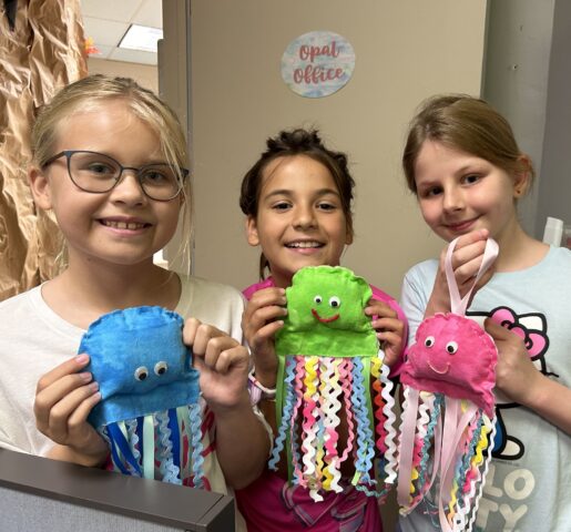 Three children smile while holding colorful jellyfish crafts with ribbon tentacles.