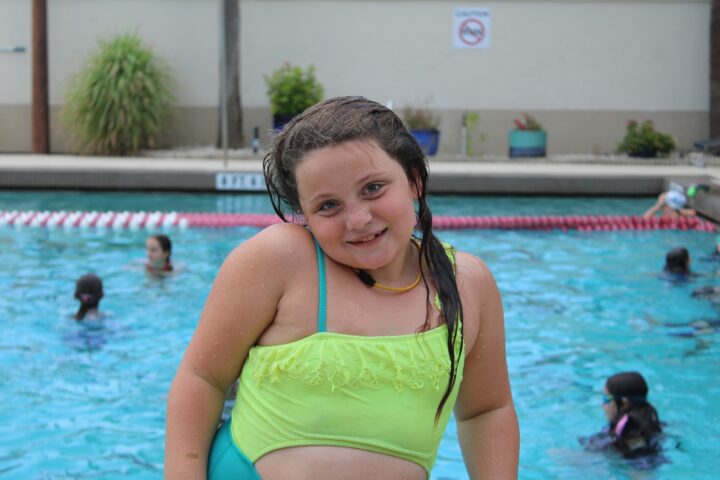 A young girl in a green swimsuit smiles at the camera while standing at the edge of a swimming pool with other swimmers in the background.