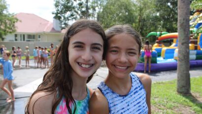 Two girls smiling at JCAmp, with a colorful inflatable slide and a group of kids in the background.