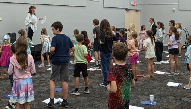 A group of children stands in a room facing an instructor holding papers. The floor is marked with papers in front of each child.