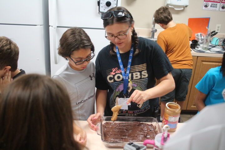 A group of people gather around a kitchen counter as a woman applies peanut butter to a chocolate cake in a baking dish.