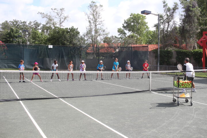 A tennis instructor guides a group of children holding rackets on a tennis court. A cart filled with tennis balls is nearby. Trees and fencing are in the background.