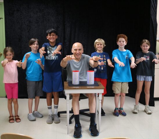 A group of children and an adult posing happily with a table and two cups in a room with a black backdrop.