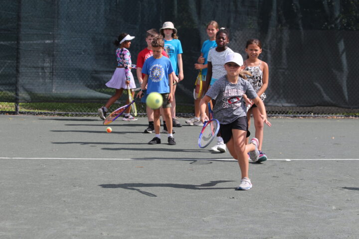 A group of children playing tennis on a court. One child is approaching a tennis ball with a racket.