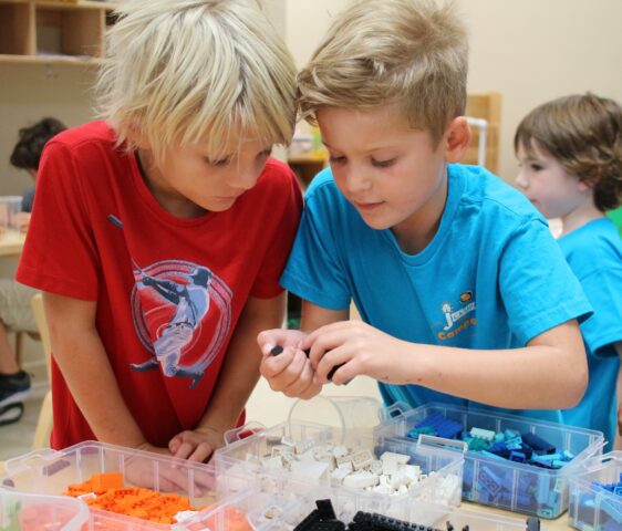 Two children in a classroom are focused on assembling small plastic pieces from a tray. Other children are visible in the background.