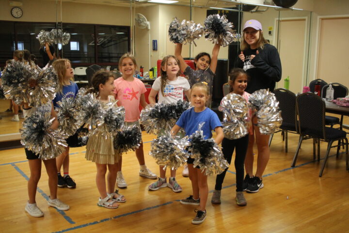 A group of young children holding silver pom-poms pose with a woman in a dance studio.