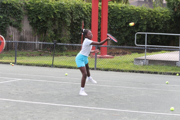 A girl playing tennis, swinging her racket to hit a ball, on an outdoor tennis court surrounded by a chain-link fence and greenery.