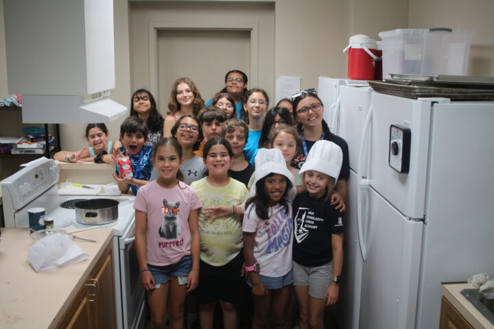 A group of children and a teenager posing in a kitchen, some wearing chef hats. They are standing near cooking appliances and ingredients.