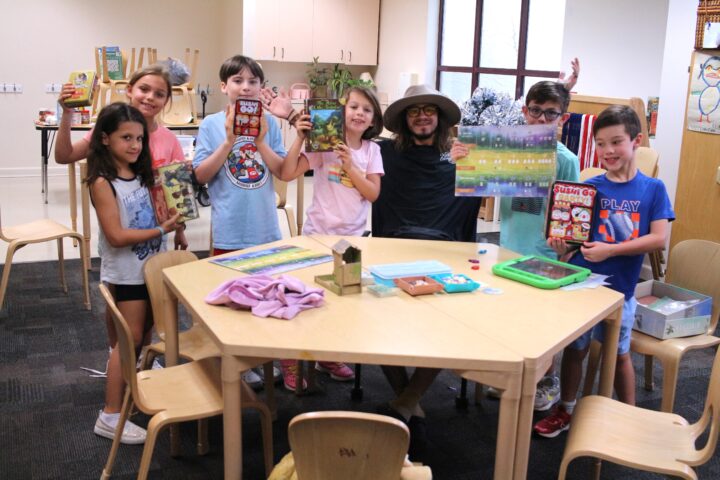 A group of children and a man gather around a table full of board games, smiling and holding up game boxes in a room with shelves and cabinets.