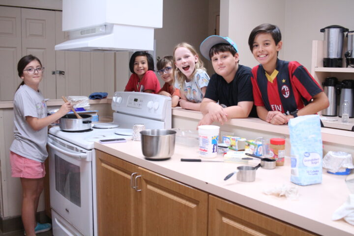 A group of kids in a kitchen, some leaning on a counter and one stirring a pot on the stove. Various baking ingredients and utensils are scattered on the counter.