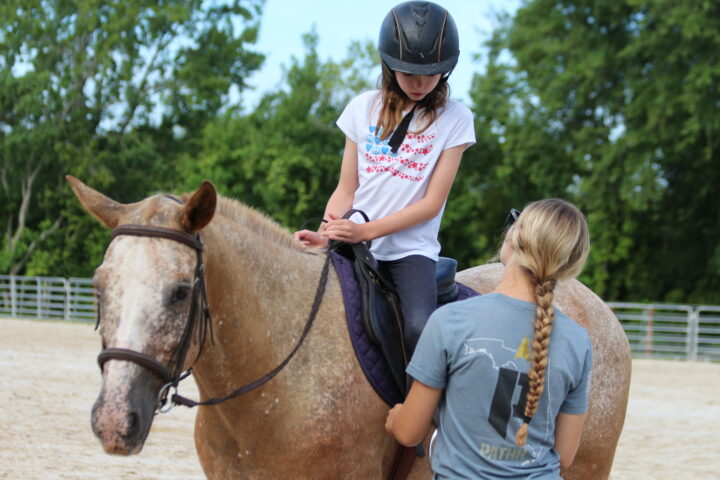 A young girl on a horse, wearing a helmet, holds the reins while an instructor with a braid and a t-shirt stands beside, outdoors in a riding arena with trees in the background.