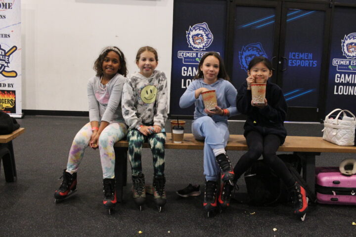 Four children in ice skates sit on a bench in an esports gaming lounge, holding snacks and smiling.