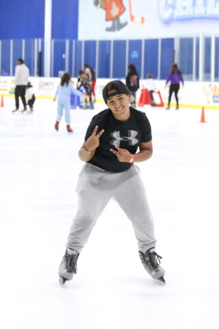 Person ice skating indoors, smiling, wearing a black cap, black shirt, and gray sweatpants, making a peace sign. Other skaters and cones are visible in the background.