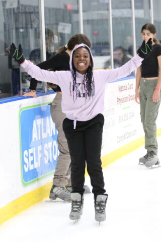 Person in a light purple hoodie and black pants ice skating indoors, smiling with arms outstretched. Others are skating in the background.