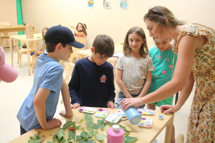 A teacher helps four children with a painting activity at a table, surrounded by art supplies and leaves.