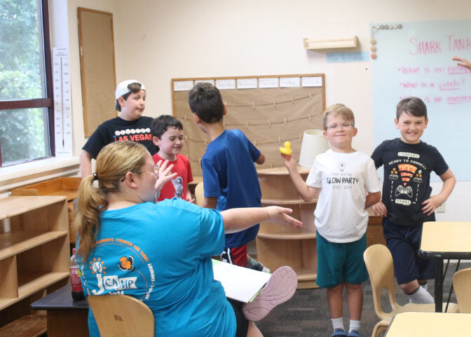 Children and an adult interact in a classroom. One child holds a rubber duck. A board with writing and a window are in the background.